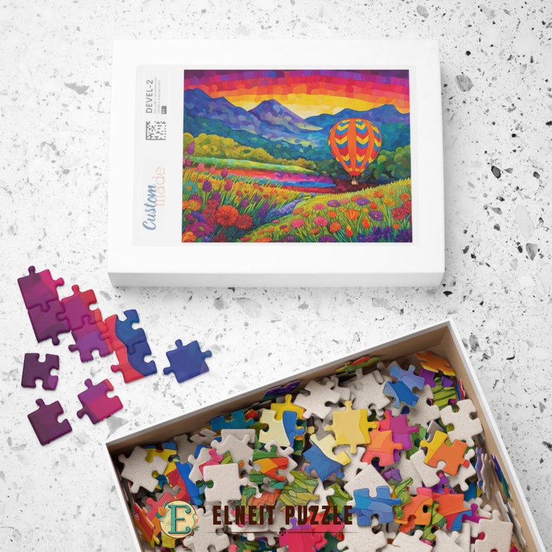 K&ouml;nnte beinhalten: Ein farbenfrohes Puzzle mit einem Hei&szlig;luftballon, der &uuml;ber einem Feld mit Blumen und Bergen im Hintergrund schwebt. Die Schachtel des Puzzles zeigt das fertige Bild des Puzzles.