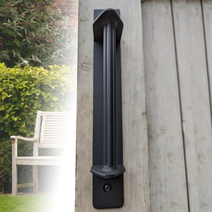 May include: Black metal door handle with a rectangular shape and a raised design. The handle is mounted on a wooden door.