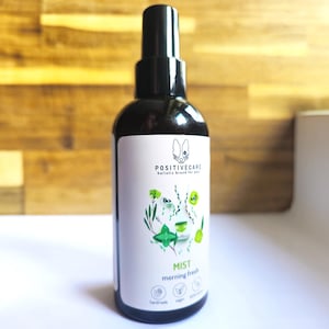 Könnte beinhalten: Eine schwarze Sprühflasche mit einem weißen Etikett. Das Etikett zeigt eine Hundeillustration und den Text "POSITIVECARE holistic brand for pets". Das Etikett lautet auch "MIST morning fresh", mit Symbolen und Text, die darauf hinweisen, dass das Produkt handgefertigt, vegan und 100% natürlich ist.