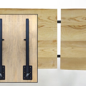 May include: Close-up of a wooden table with a light finish, featuring two black metal folding table leg supports. The supports are attached to the underside of the table with screws. The table is made of wood.