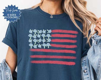 May include: Navy blue t-shirt featuring a floral American flag design. The flag's stripes are red, and the stars are replaced with blue flowers. The shirt has the text "Pick from 6 sizes 5 colors" in a blue starburst.