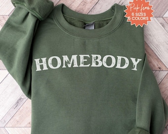 May include: A military green sweatshirt with the word "HOMEBODY" printed in white. The sweatshirt has a crew neck and long sleeves. The image also includes text that says "Pick from 6 sizes 5 colors" and "Military Green".