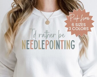 May include: White sweatshirt with the text "I'd rather be needlepointing" in colorful letters. The sweatshirt is being modeled by a person. A starburst sticker with the text "Pick from 6 sizes 3 colors" is in the top right corner of the image.
