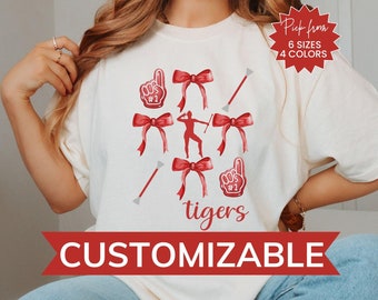 May include: A white t-shirt with a red and white design featuring a cheerleader, bows, and the words "tigers". The text "Pick from 6 sizes 4 colors" and "CUSTOMIZABLE" are also visible on the shirt.