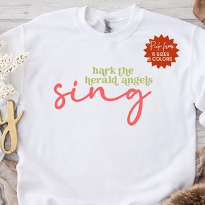 May include: White sweatshirt with the text "hark the herald angels sing" in red lettering. The text is in a cursive font. A red starburst sticker with the text "Pick from 6 sizes 5 colors" is in the upper right corner of the image.
