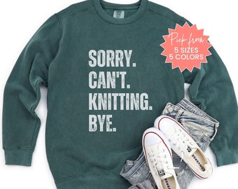 May include: A dark green sweatshirt with the text "SORRY. CAN'T. KNITTING. BYE." in white distressed lettering. The sweatshirt is shown on a model with blue jeans and white sneakers. The text "Pick from 5 sizes 5 colors" is on a red starburst.