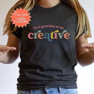 May include: A dark grey t-shirt with the text "it's a good day to be creative" in colorful letters. The text is in a retro font style.