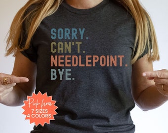May include: A dark gray t-shirt with the text "SORRY. CAN'T. NEEDLEPOINT. BYE." in a vintage style font. The text is in shades of blue, green, and orange. The shirt is being worn by a person with long blonde hair. The text "Pick from 7 sizes 4 colors" is on a red starburst sticker.