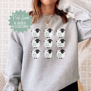May include: A gray sweatshirt with a pattern of nine white sheep with black legs and a green star design on their bodies. The text "Pick from 6 sizes 4 colors" is on a starburst sticker.
