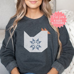 May include: Dark gray sweatshirt featuring a geometric chicken design with a star pattern. The chicken is white with blue and orange accents. The image includes text that says "Pick from 6 sizes 5 colors".