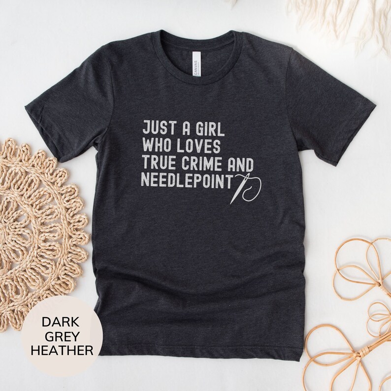 May include: Dark heather grey t-shirt with the text "Just a girl who loves true crime and needlepoint" and a stylized needle and thread design.