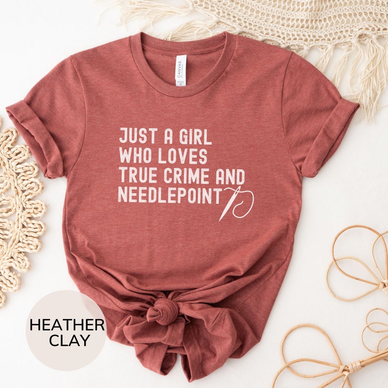 May include: A heather clay t-shirt with the text "Just a girl who loves true crime and needlepoint" and a stylized needle and thread graphic.