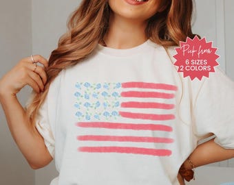 May include: Cream-colored t-shirt featuring a watercolor American flag design. The flag's stars are replaced with blue floral patterns, and the stripes are a soft red. The shirt also has the text "Pick from 6 sizes 2 colors".