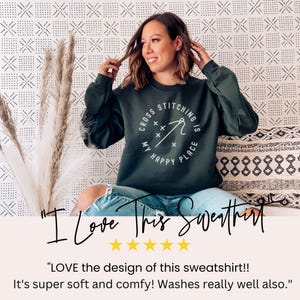 May include: Dark green sweatshirt with the text "Cross Stitching is My Happy Place" in a white circular design. The sweatshirt is long-sleeved and worn by a person. The image includes text that says "I Love This Sweatshirt".