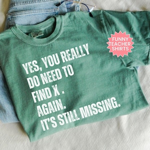 May include: A sage green t-shirt with the text "YES, YOU REALLY DO NEED TO FIND X. AGAIN. IT'S STILL MISSING." in white. A pink starburst graphic reads "FUNNY TEACHER SHIRTS". The shirt is folded on top of blue jeans.