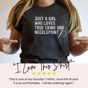 May include: A dark gray t-shirt with the text "Just a girl who loves true crime and needlepoint" in white. A white needle and thread design is next to the text. The text "I Love This Shirt" is written in black cursive with five gold stars below it.