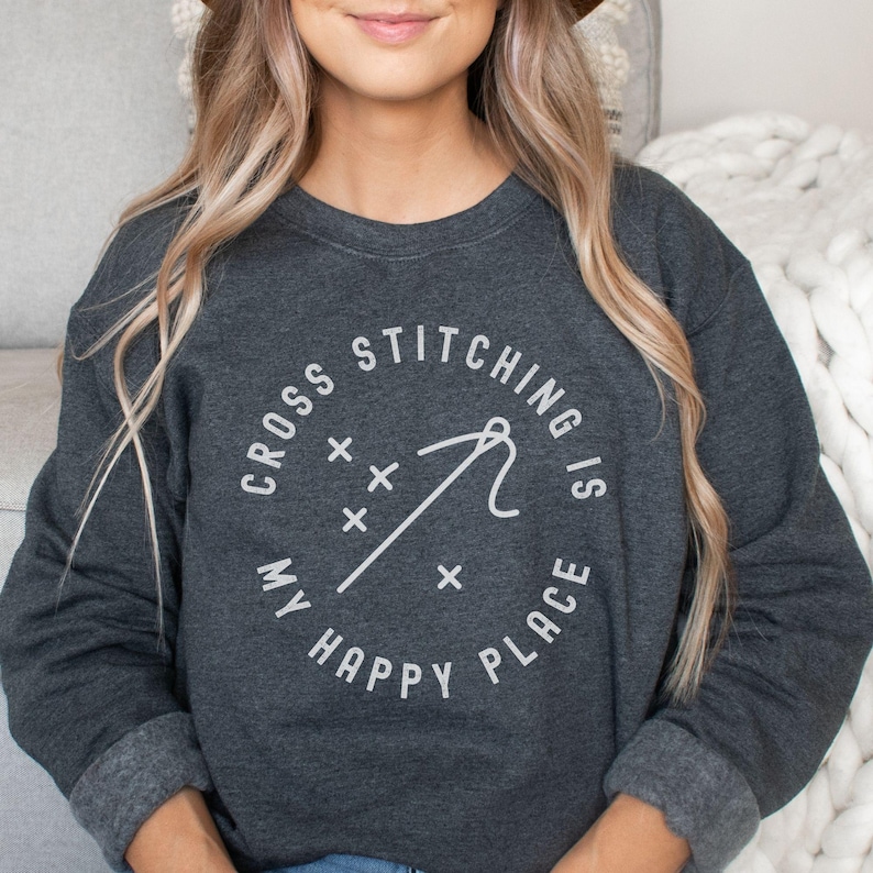 May include: Dark gray sweatshirt with the text "CROSS STITCHING IS MY HAPPY PLACE" in a white circular design. The design includes a needle, thread, and cross stitch symbols. The sweatshirt has a crew neck and long sleeves.