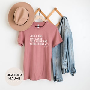 May include: A heather mauve t-shirt with the text "Just a girl who loves true crime and needlepoint" in white. The shirt is hanging on a wooden hanger.