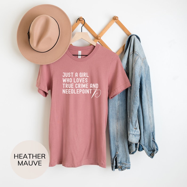 May include: A heather mauve t-shirt with the text "Just a girl who loves true crime and needlepoint" in white. The shirt is hanging on a wooden hanger.