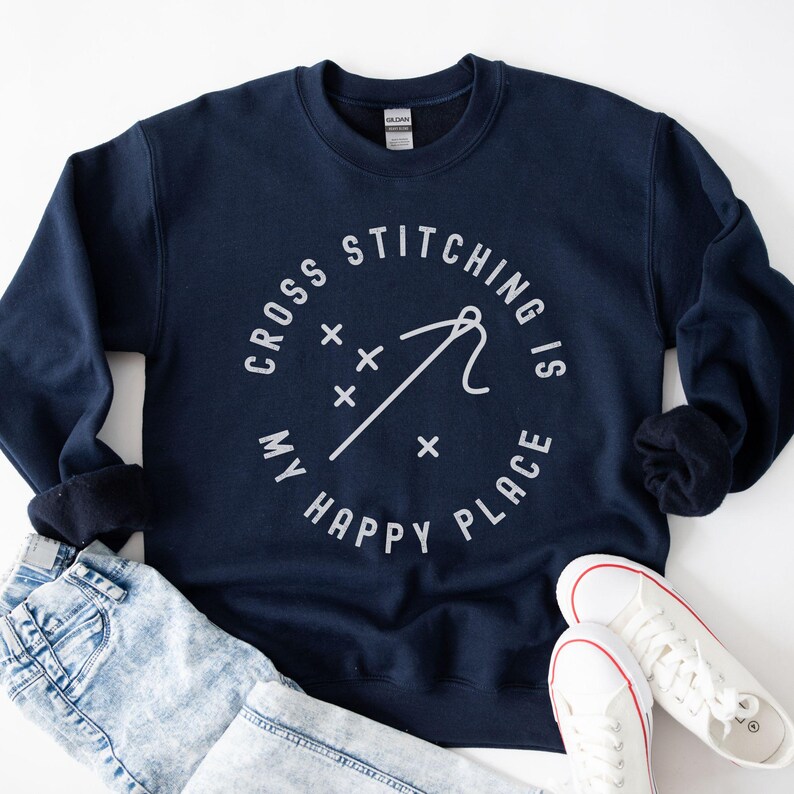 May include: Navy blue sweatshirt with the text "CROSS STITCHING IS MY HAPPY PLACE" in a circular design. The design includes a needle and thread graphic. The sweatshirt is paired with light wash jeans and white sneakers.