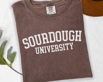 May include: A brown t-shirt with the words "SOURDOUGH UNIVERSITY" printed in white block letters. The shirt has a crew neck and short sleeves. The Comfort Colors tag is visible at the neckline. A brown hat is partially visible in the background.
