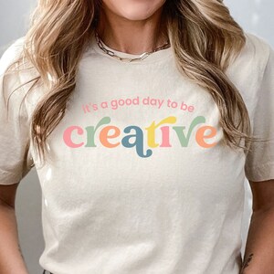 May include: A beige t-shirt with the text "It's a good day to be creative" in colorful letters. The shirt is being worn by a person with long blonde hair. The text "COLOR - NATURAL" is visible at the bottom of the image.