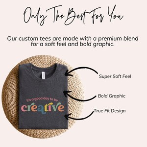 May include: A dark gray t-shirt with the text "it's a good day to be creative" printed in colorful letters. The shirt is folded and displayed on a woven mat. The text "Super Soft Feel", "Bold Graphic", and "True Fit Design" are written next to the shirt.