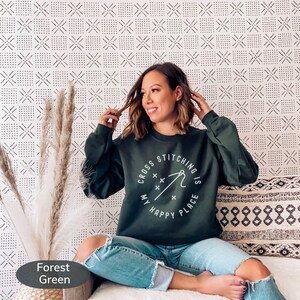 May include: Forest green sweatshirt with the text "Cross Stitching is My Happy Place" in a white circular design. The sweatshirt is paired with blue jeans. The background features a patterned wall and decorative pillows.