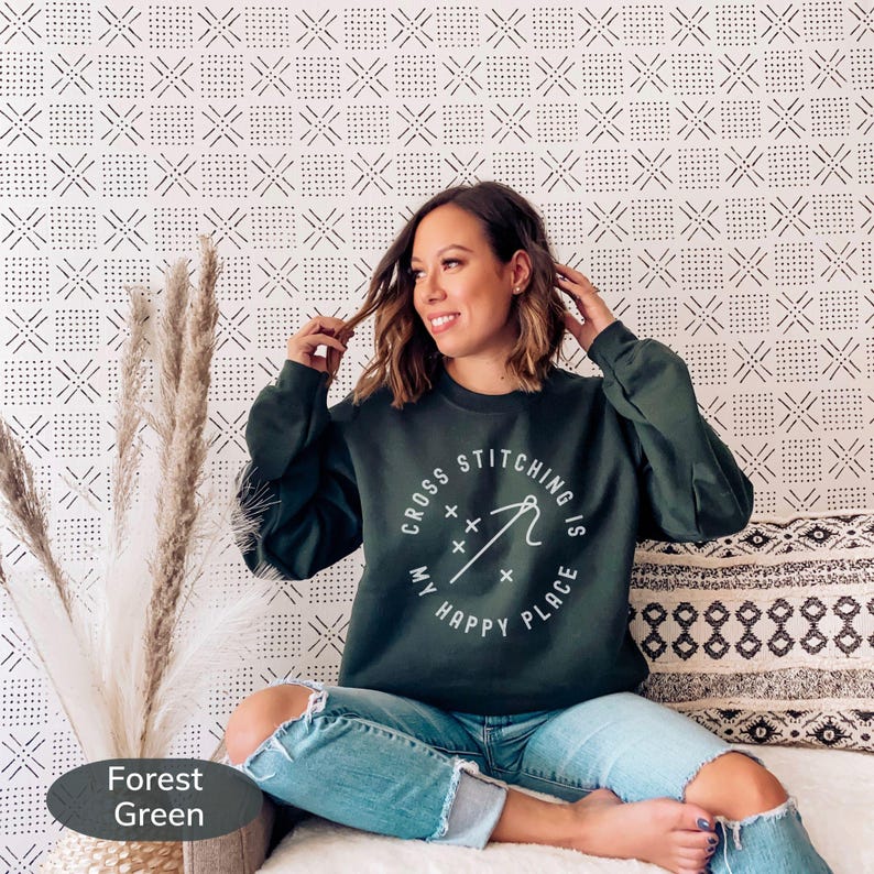May include: Forest green sweatshirt with the text "Cross Stitching is My Happy Place" in a white circular design. The sweatshirt is paired with blue jeans. The background features a patterned wall and decorative pillows.
