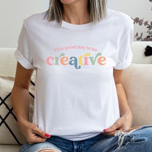 May include: White t-shirt with the text "It's a good day to be creative" in colorful letters.