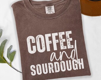 May include: A brown t-shirt with the words "COFFEE and SOURDOUGH" printed in white. The shirt is folded and has a tag that says "COMFORT COLORS". A brown hat is visible in the background.