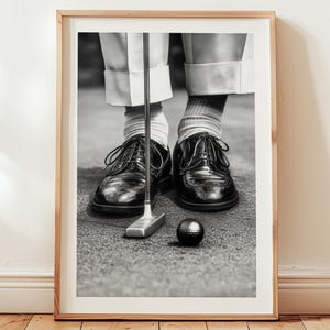 May include: Black and white framed print of a golfer putting. The image shows the golfer's feet, black leather shoes, golf club, and a golf ball on the green. The golfer is wearing white pants and striped socks.