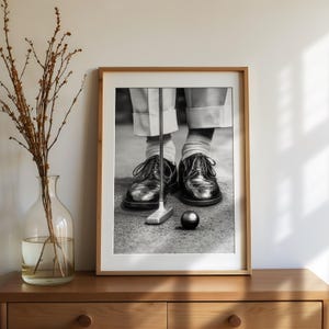 May include: Black and white framed print of a golfer putting. The image shows the golfer's feet in black leather shoes, white socks, and light-colored pants. A golf club and ball are in focus.