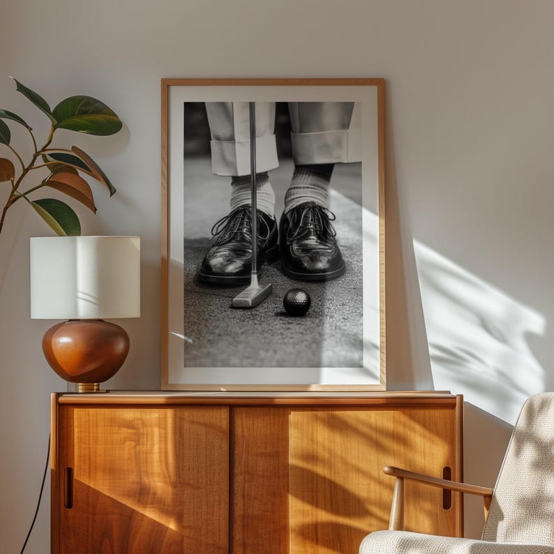 May include: Black and white framed print of a golfer's feet, shoes, and golf club. The golfer is wearing black leather shoes, white socks, and light-colored pants. A golf ball and putter are on the ground.
