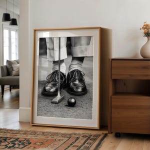 May include: Black and white framed print of a golfer putting. The image shows the golfer's feet, black leather shoes, golf club, and a golf ball. The frame is light wood.