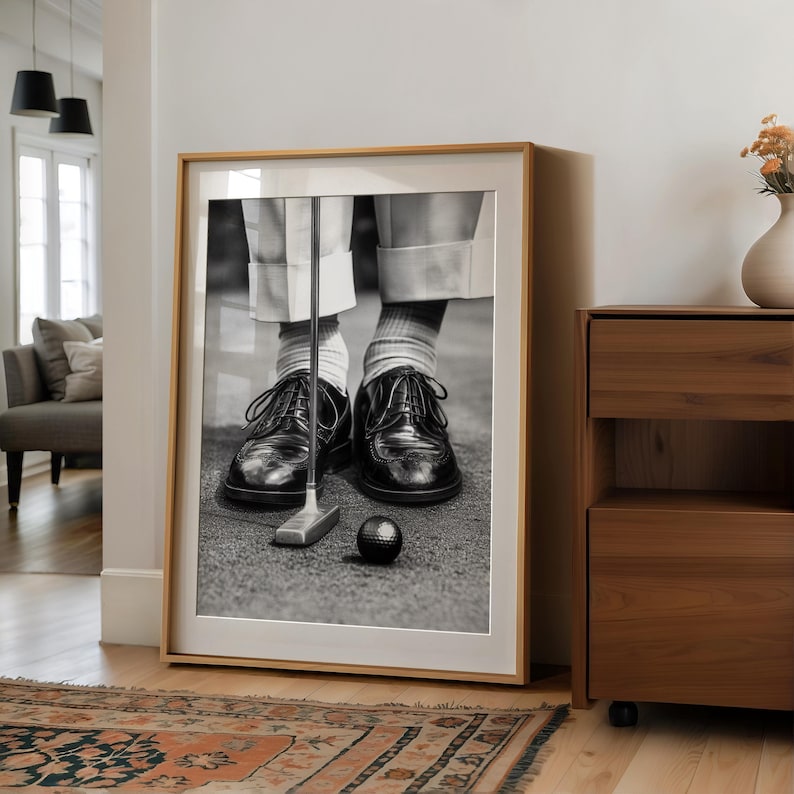 May include: Black and white framed print of a golfer putting. The image shows the golfer's feet, black leather shoes, golf club, and a golf ball. The frame is light wood.
