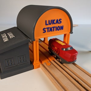 May include: A red toy train with a yellow fuel tank is passing through a grey and orange toy train station with the name "Lukas Station" printed on the front.