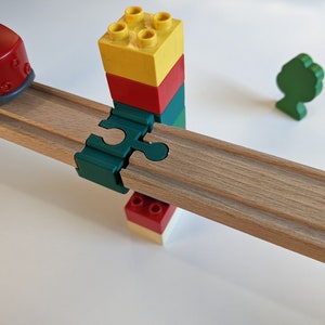 Könnte beinhalten: Ein roter Spielzeugzug auf einem Holzgleis mit einem grünen und roten Blockturm rechts. Das Gleis wird von einem grünen Kunststoff-Verbindungsstück getragen.