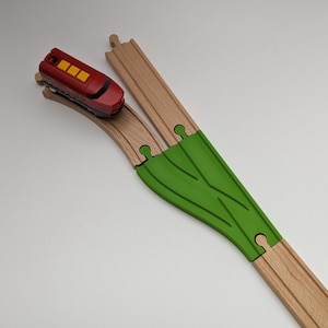 Op de afbeelding: Een rode speelgoedtrein met gele accenten bevindt zich op een houten spoor met een groene plastic wissel. De trein rijdt een bocht op het spoor.