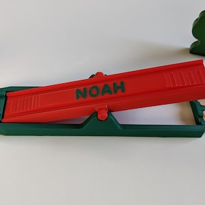 Op de afbeelding: Een rode houten treinbaanhelling met de naam "NOAH" erop gedrukt. De helling is bevestigd aan een groene basis en is in een hoek omhoog gekanteld.