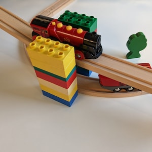 Op de afbeelding: Een rode speelgoedtrein met een zwarte locomotief en gele wielen rijdt op een houten spoor. De trein rijdt over een brug gemaakt van houten planken. Aan beide kanten van het spoor zijn kleurrijke bouwstenen gestapeld.