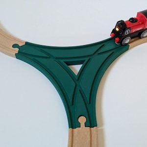 Puede incluir: Una vía de tren de madera verde con un tren de juguete rojo sobre ella. La vía es un cambio de tres vías, que permite al tren ir en tres direcciones diferentes.