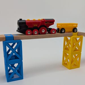 Può includere: Un locomotore giocattolo rosso e nero con un vagone giocattolo giallo che attraversa un ponte di legno sostenuto da blocchi blu e gialli. Il motore ha il numero 529 stampato sul lato.