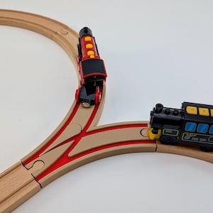 Op de afbeelding: Een rode en zwarte speelgoedtrein met gele accenten bevindt zich op een houten spoor. De locomotief bevindt zich op een gebogen spoor dat splitst in twee sporen. Een zwarte en gele speelgoedtrein bevindt zich op het andere spoor.