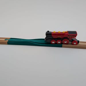 Op de afbeelding: Een rode en zwarte speelgoedtrein met gele accenten en het nummer 529 aan de zijkant, die over een groen houten spoor rijdt.