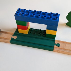 Op de afbeelding: Een speelgoedtreinset met een rode speelgoedtrein, een groene houten boom en een blauwe, groene, rode en gele Lego-brug.