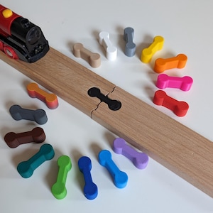 Può includere: Un set di connettori per binari del treno in legno colorati, tra cui rosso, arancione, giallo, verde, blu, viola, marrone, grigio e bianco. I connettori sono disposti in un cerchio attorno a un binario del treno in legno.