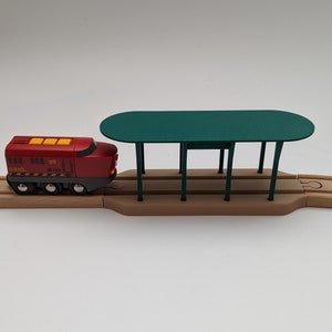 Könnte beinhalten: Eine rote Spielzeug-Lokomotive mit der Aufschrift "BRIO" und der Nummer "JR 103" auf der Seite, die in einen grünen Spielzeug-Bahnhof mit einer Holzplattform einfährt.