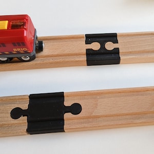 Könnte beinhalten: Eine rote Spielzeug-Lokomotive mit gelben Fenstern und einem schwarzen BRIO-Logo an der Seite, die auf einem hölzernen Gleis mit schwarzen Kunststoff-Gleisverbindern fährt.