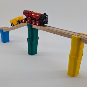 May include: A red and black toy train with a yellow toy car on a wooden track bridge with green, blue, and yellow supports.